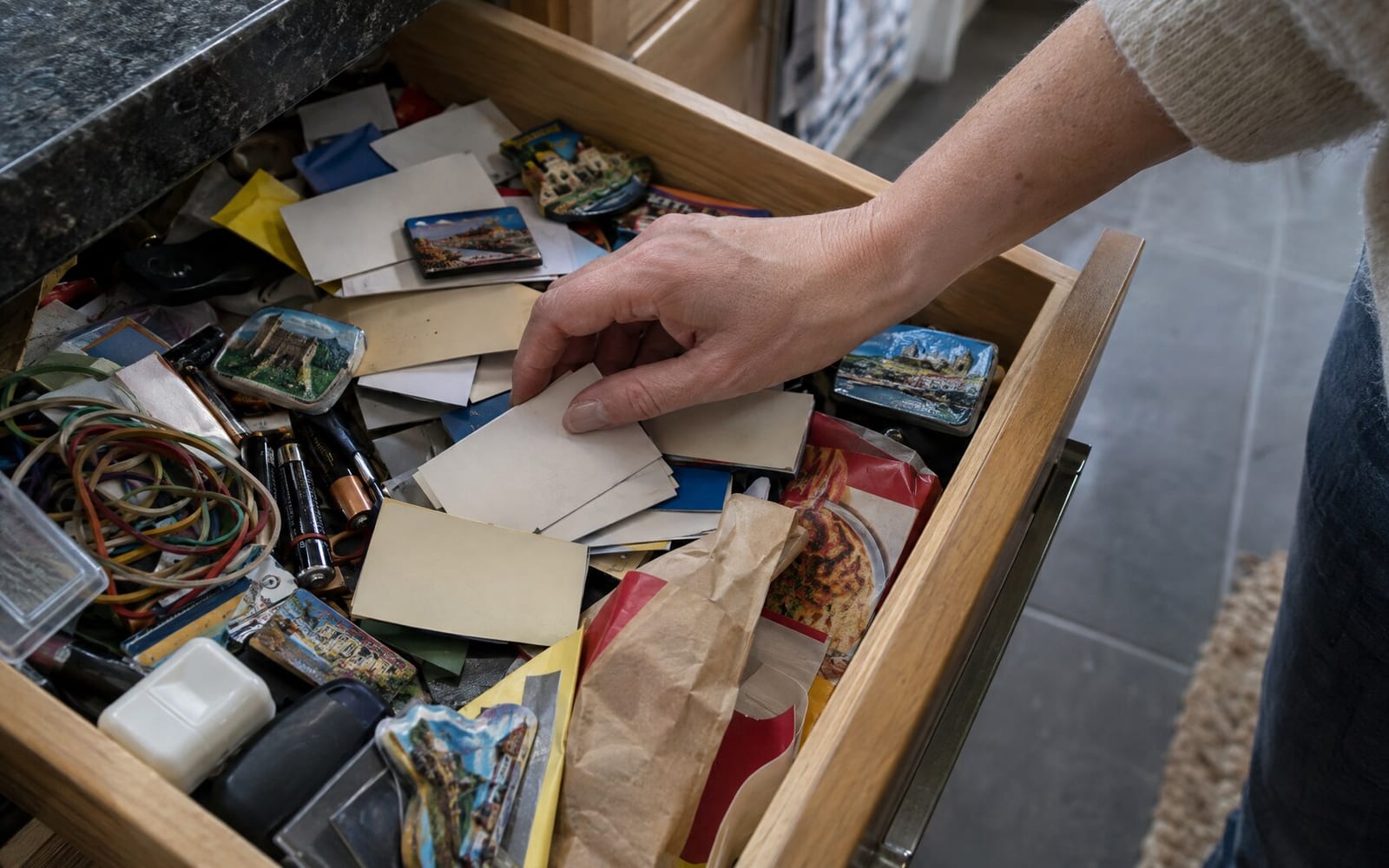 A hand rummaging through a kitchen drawer full of old paper business cards, fridge magnets and takeaway menus