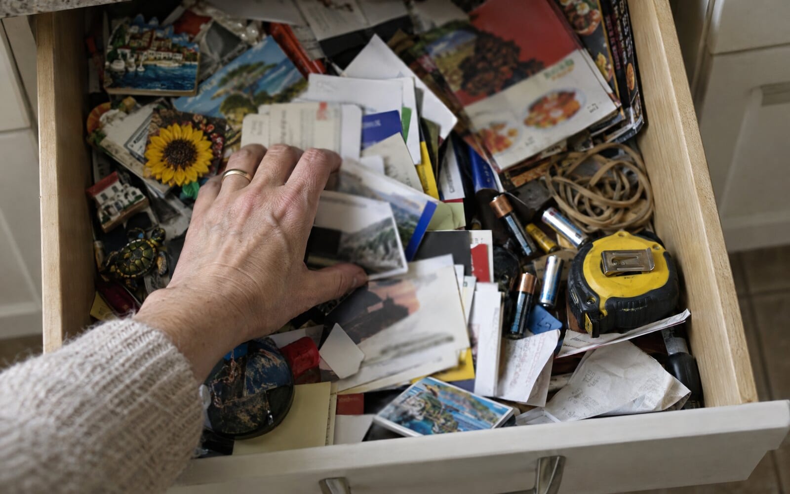An open kitchen drawer full of paper business cards, fridge magnets and takeaway menus, with a hand searching through it